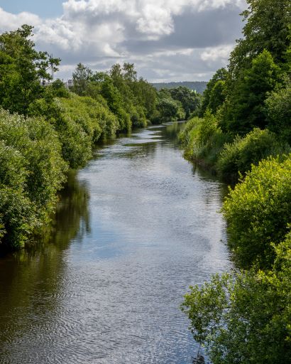 A river with lush green trees on both sides. Sky with clouds reflects in water.  Flows through Partille center, nature in the middle of the city.