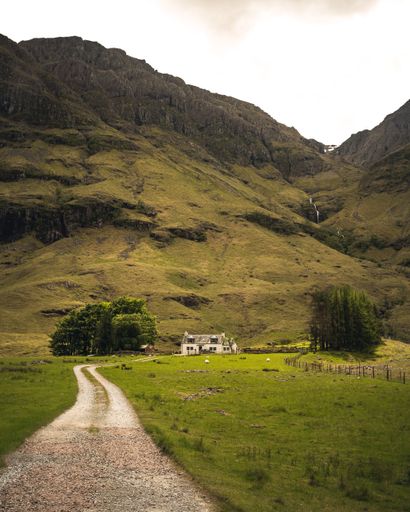 A vertical shot of a road to a small farmhouse under high cliff hills in Glencoe Village in Scotland