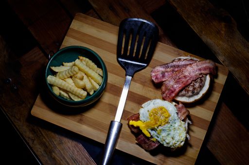 A high angle closeup shot of a breakfast of bacon, fries, and egg next to a spatula on a wooden cutting board
