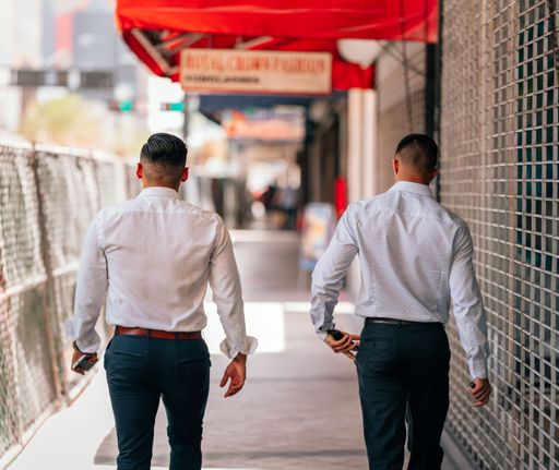 A back view of two men wearing white shirt and walking in footpath surrounded by metal fences