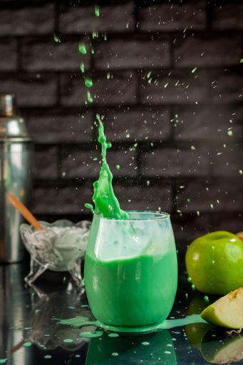 A vertical shot of a green apple smoothie juice with splashes on a black table and apples in the background