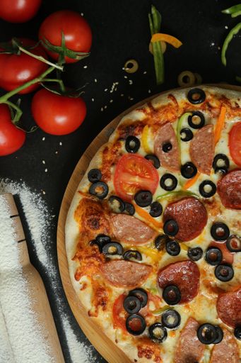 A vertical top view of a sliced pizza with olives with black background and fresh vegetables