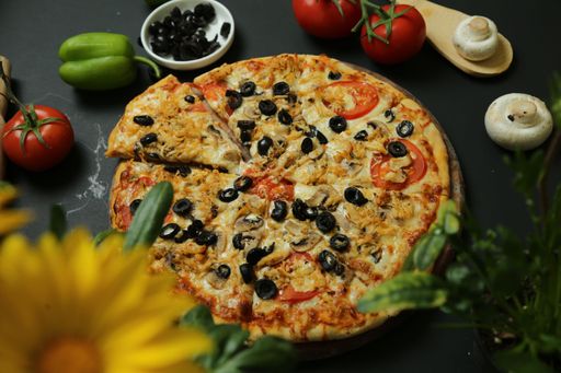 A close-up shot of a sliced pizza with olives with black background surrounded by fresh vegetables