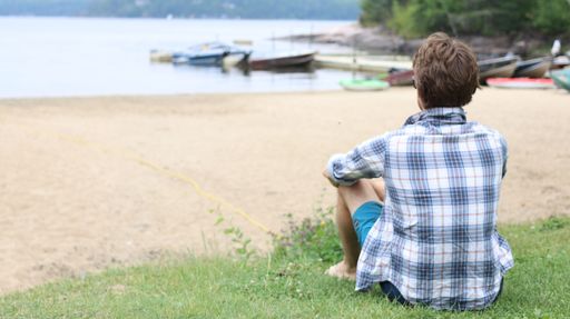 A Caucasian man seen from behind, sitting on green grass on a beach, watching the boat docks