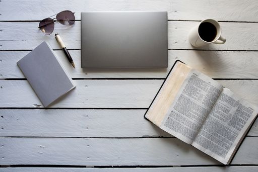 An overhead shot of an open bible near a laptop, coffee and a notebook with a pen on a white wooden surface