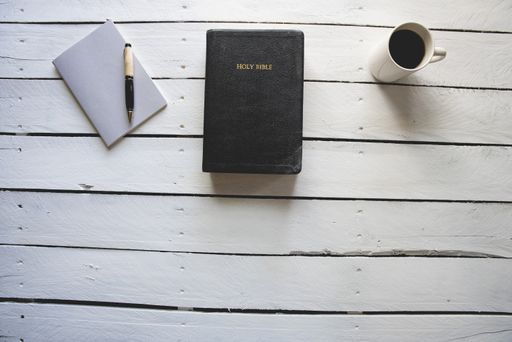 An overhead shot of the holy bible in between a coffee and a notepad with a pen on a white wooden surface