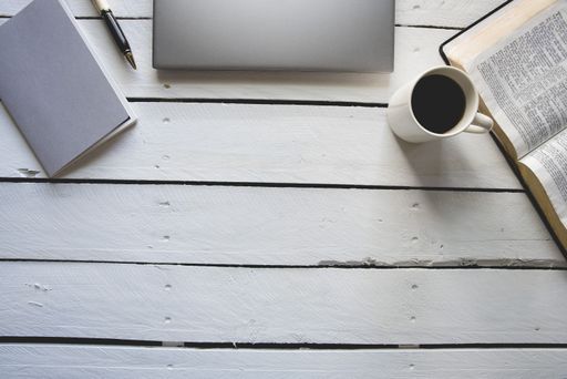 An overhead shot of a white wooden surface with a laptop, bible, coffee and a notepad with a pen on the top