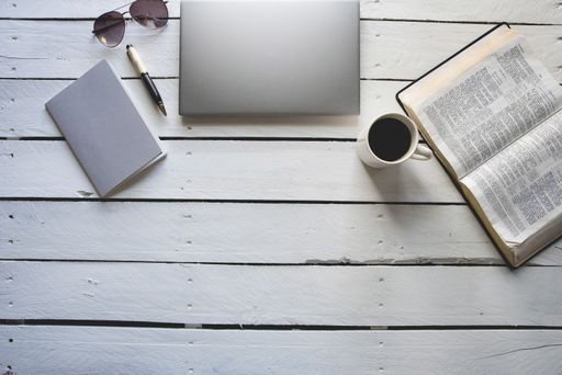 An overhead shot of an open bible near a coffee and, laptob, notepad with a pen on a white wooden surface
