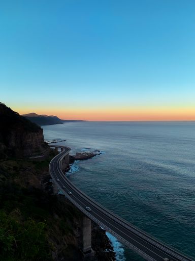 A beautiful shot of cars on a curly bridge road with sea view and mountain during sunset