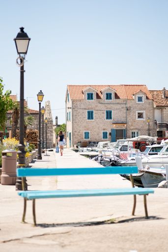 A vertical shot of a blue bench with a girl walking in the background at the docks on Island Prvic, Croatia