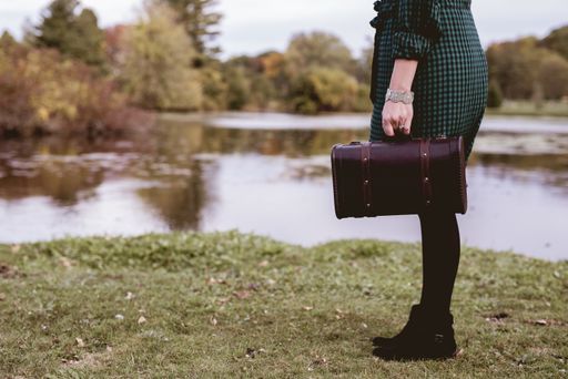 A closeup shot of a female standing in a grassy field and holding an old suitcase with a blurred background