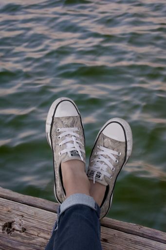 A vertical shot of a woman's feet in gray Converse shoes relaxing on the wooden pier by the lake