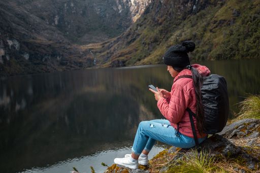 A Caucasian girl in a pink jacket with a backpack sitting on a rock by the lake and looking at her phone in the mountains