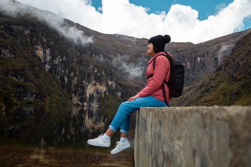 A smiling girl in a pink jacket with a backpack sitting by the lake in the mountains and looking at nature