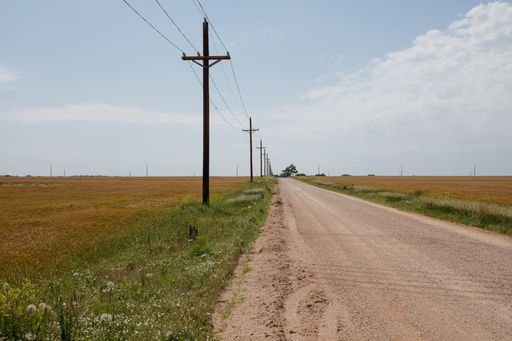 An empty road along electric poles in the middle of a field under a blue sky