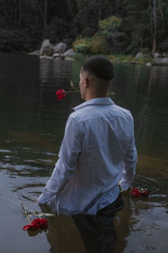 A vertical shot of a young man standing in a river with a red rose in his teeth and a reflection on the water surface