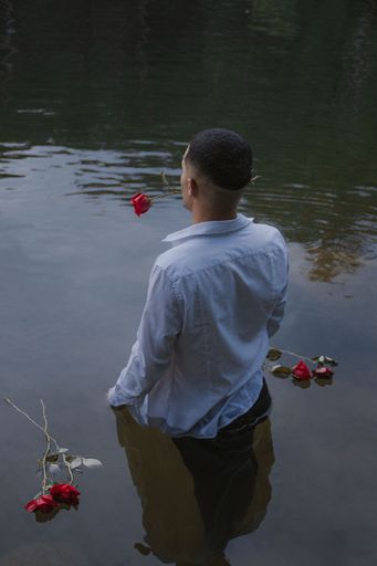 A vertical shot of a young man standing in a river with a red rose in his teeth and a reflection on the water surface