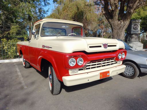 Old red and cream utility pickup truck Ford F 100 Styleside bed 1960 in a park. Front view. Grille. Nature, trees. Classic car show