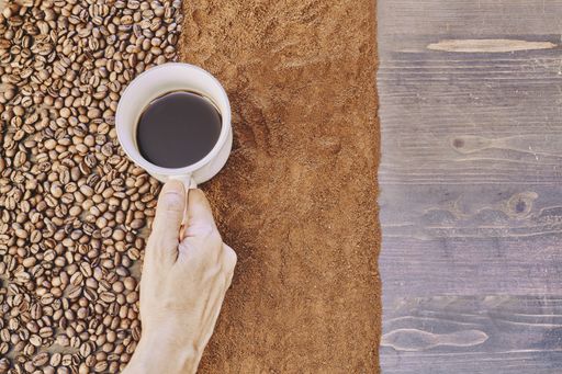 A top view of a person holding a cup of coffee with roasted and ground coffee beans on the table