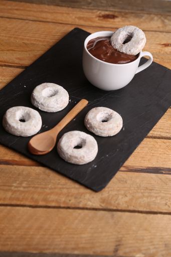 A vertical shot of homemade bagels, a cup of hot chocolate, and a wooden spoon on a black sheet on a wooden surface