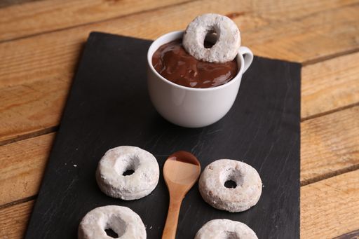 A closeup of homemade bagels, a cup of hot chocolate, and a wooden spoon on a black sheet, on a wooden surface