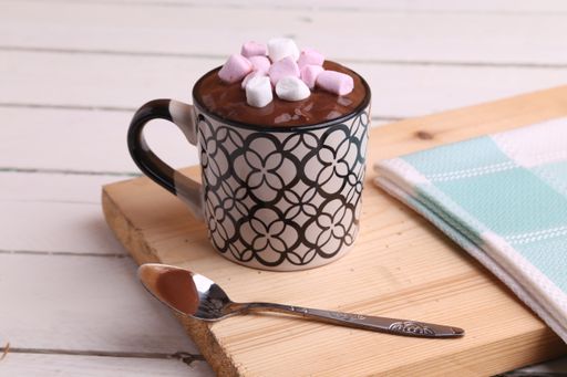 A closeup shot of a cup of hot chocolate with marshmallows on a wooden board with a spoon and a towel next to it