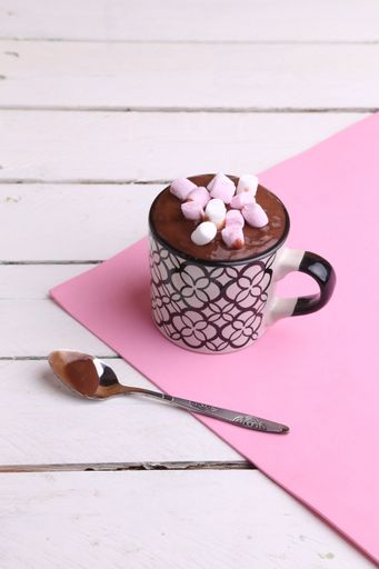 A vertical shot of thick hot chocolate in a patterned mug topped with marshmallows with a spoon on a pink sheet, on a white wooden surface