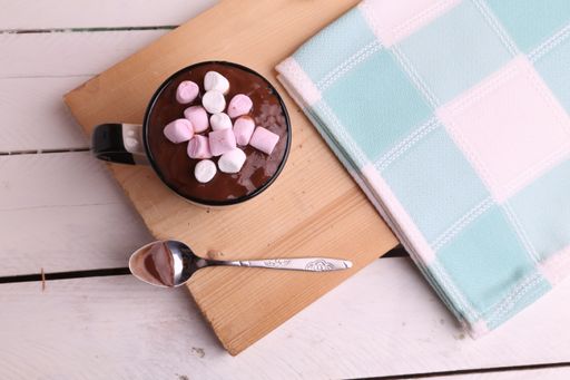 A flat lay shot of a cup of hot chocolate with marshmallows on a wooden board with a spoon and a towel next to it