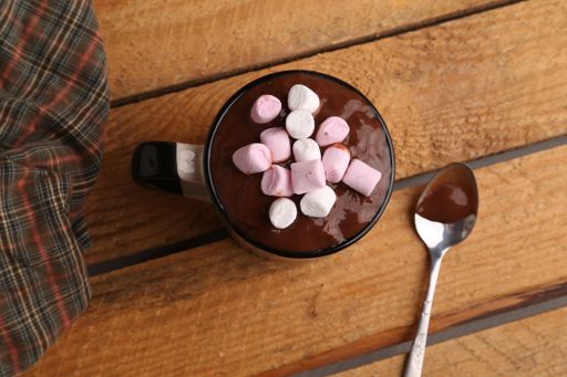 A flat lay shot of a mug of hot chocolate with marshmallows on and a spoon on a wooden table