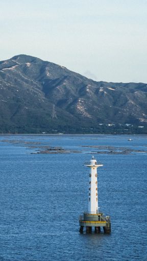 A vertical shot of a lighthouse in the beautiful blue sea with mountains in the background