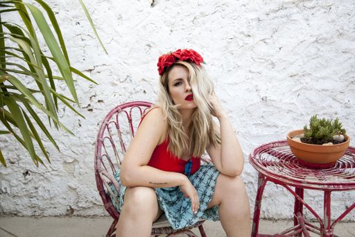 A young female with white hair and a red floral headband posing while sitting on a chair near a potted palm in front of a white grunge wall