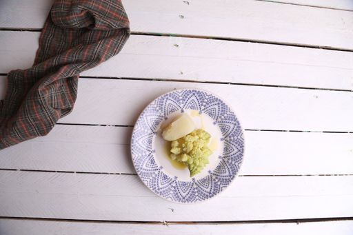 A flat lay shot of boiled broccoli and potato on a plate and a towel on a white wooden table