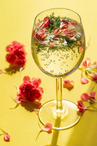 A vertical shot of a drink in a wineglass with flower petals and dill on a yellow background