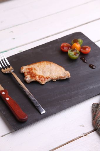 A vertical shot of a meat piece, a knife and a fork and small tomatoes on a black wooden board