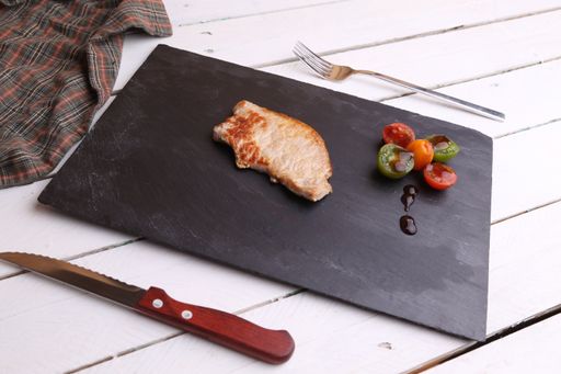 A black wooden board with pork chop steak next to a knife and fork