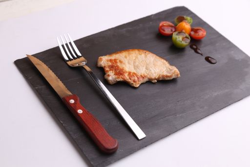 A high angle shot of grilled chicken, a fork, a knife and colorful Romain tomatoes on a black kitchen board