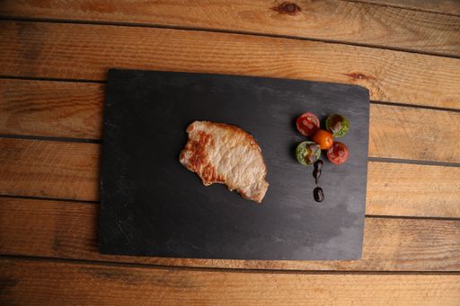 A top view of a pork tenderloin steak next to rainbow cherry tomatoes on a black plate, on a wooden surface