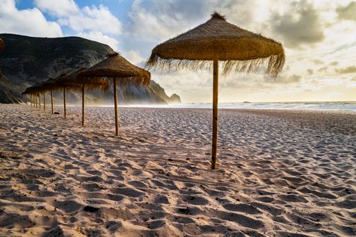 The straw parasols on a sandy beach under a partly cloudy sky.