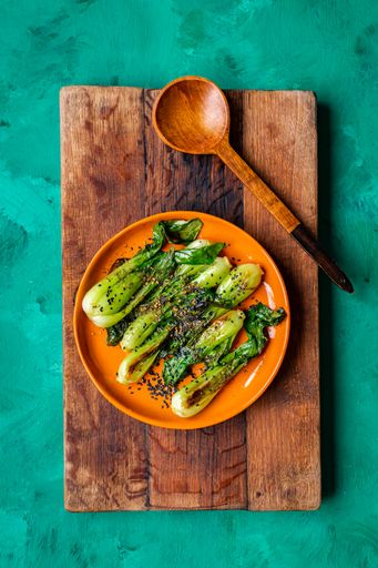 Grilled pak choi (Asian greens) with sesame seed dressing on orange plate on wooden board and green surface with wooden serving spoon