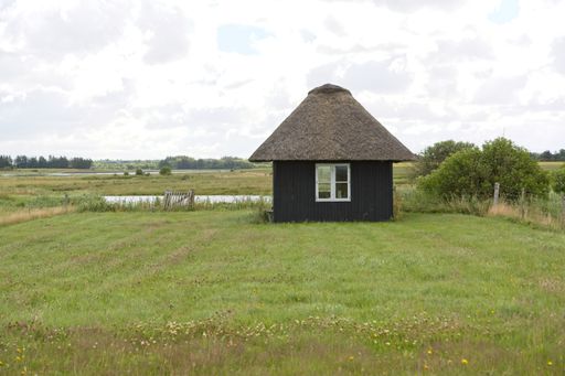 A small wooden cabin in a green grassy field under a cloudy sky