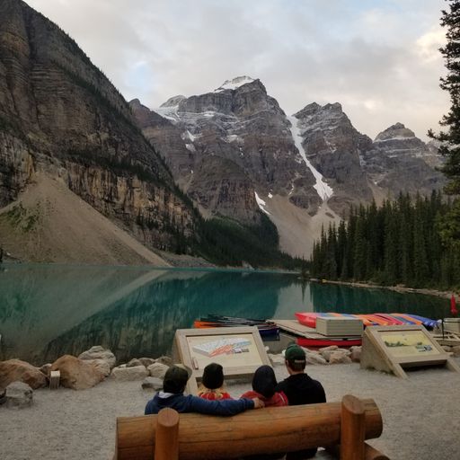 A view of people sitting on wooden bench near Moraine Lake surrounded by snow covered mountains