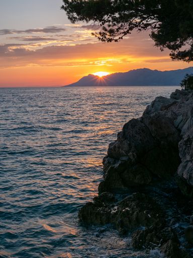 A vertical view from the rocky shore of the bright sun in orange sunset sky behind the mountain