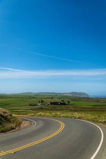 A vertical shot of a two-way asphalt road passing through green fields under a blue sky