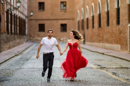 A Caucasian male holding hands with a Caucasian female in a beautiful red dress, walking on the background of buildings