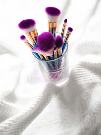 A vertical closeup shot of numerous purple and blue makeup brushes in a glass brush holder on a white cloth