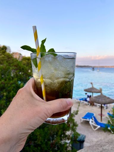 A vertical closeup shot of a hand holding a Caipirinha drink with the Illetas beach in the background