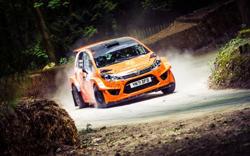 A white orange car racing on the road through a green forest at the Goodwood Festival of Speed