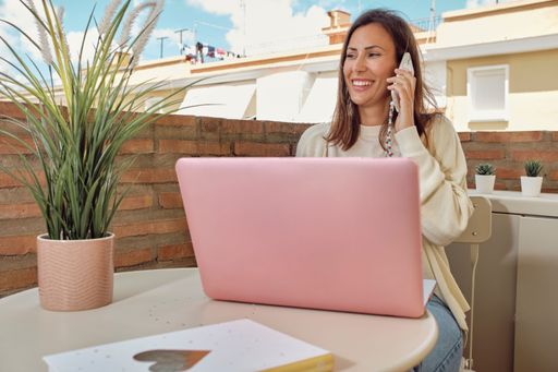 A woman working on a laptop while sitting in the balcony and talking on the phone on a sunny day