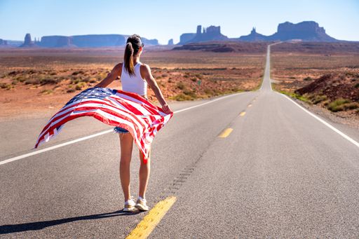 A brunette girl walking on an empty road holding the United States flag in the Monument Valley