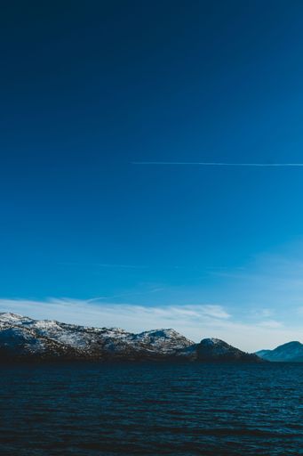 A vertical shot of an Okanagan lake in winter with snow-capped mountains and blue sky with a visible plane trail